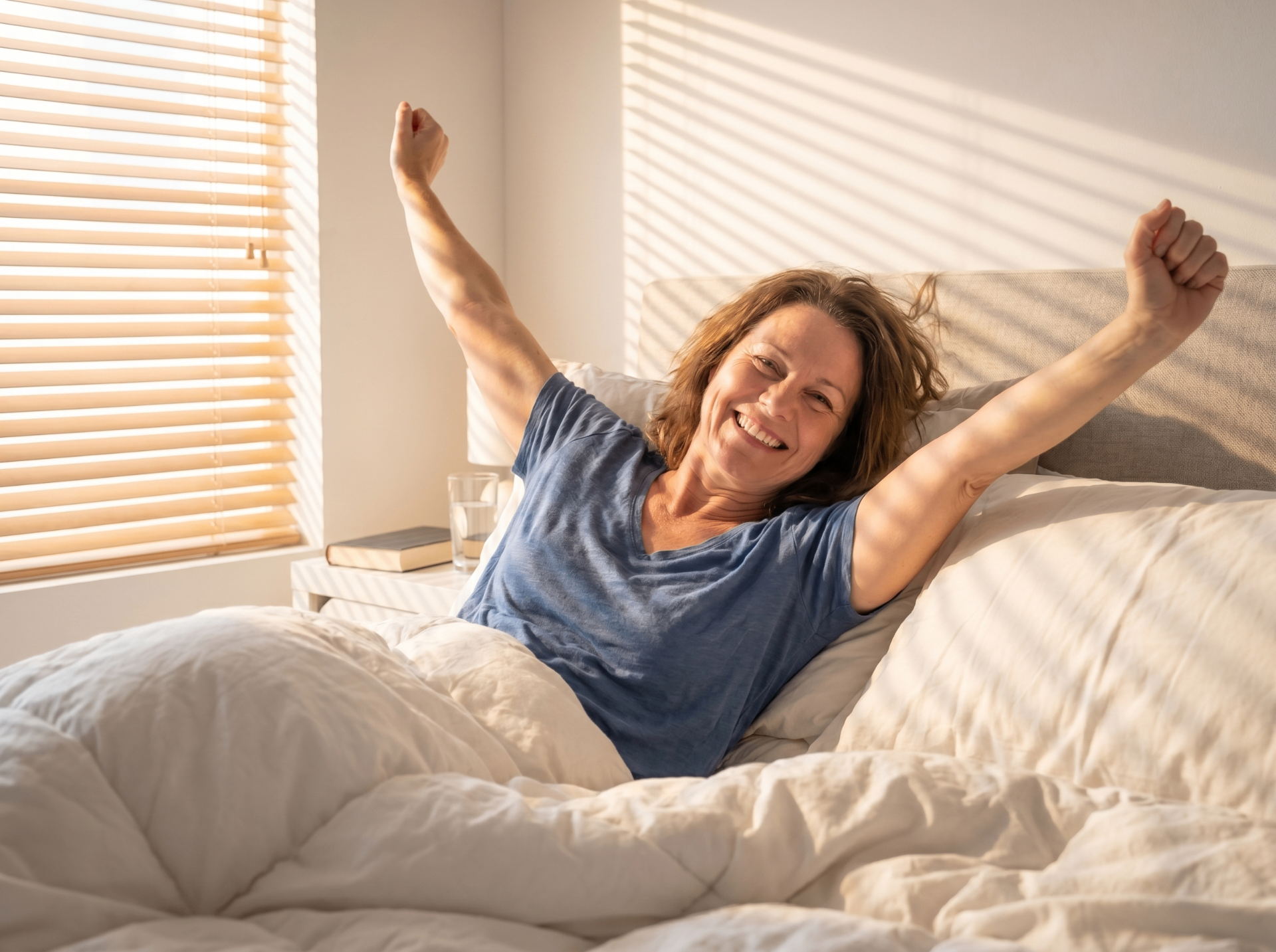 A woman waking up rested and stretching in a sunlit bedroom